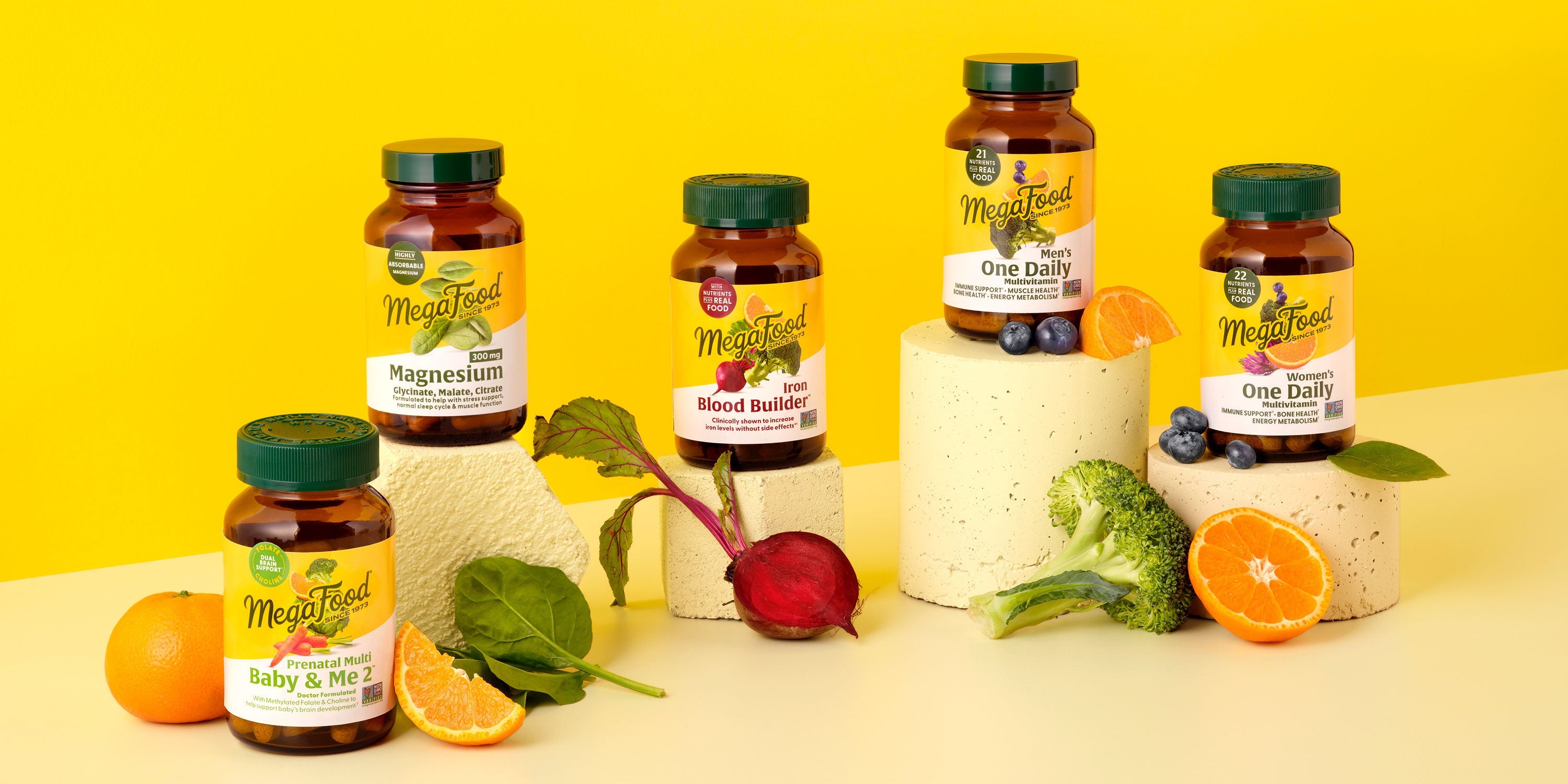 Five MegaFood supplement bottles are arranged on cream blocks, surrounded by fresh fruits and vegetables like broccoli, beets, orange, blueberries, and spinach, set against a bright yellow background.