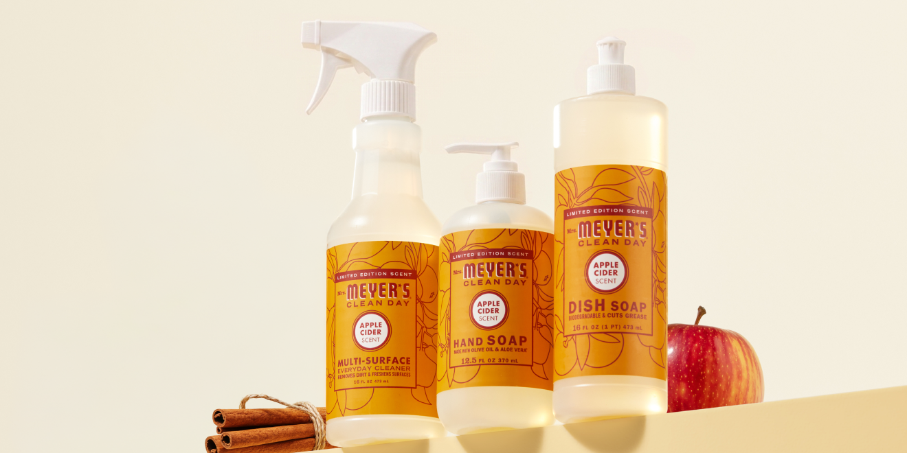 Mrs. Meyers cleaning products with autumn-themed labels on an orange background, surrounded by a candle, flowers, cinnamon sticks, acorns, and a red maple leaf.