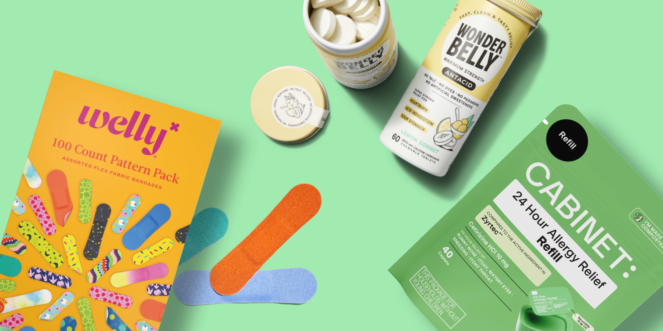 A flat lay of wellness products on a green background, including Welly bandages, loose bandages, a pill bottle, Wonderbelly antacid tablets, and Cabinet allergy relief tablets.