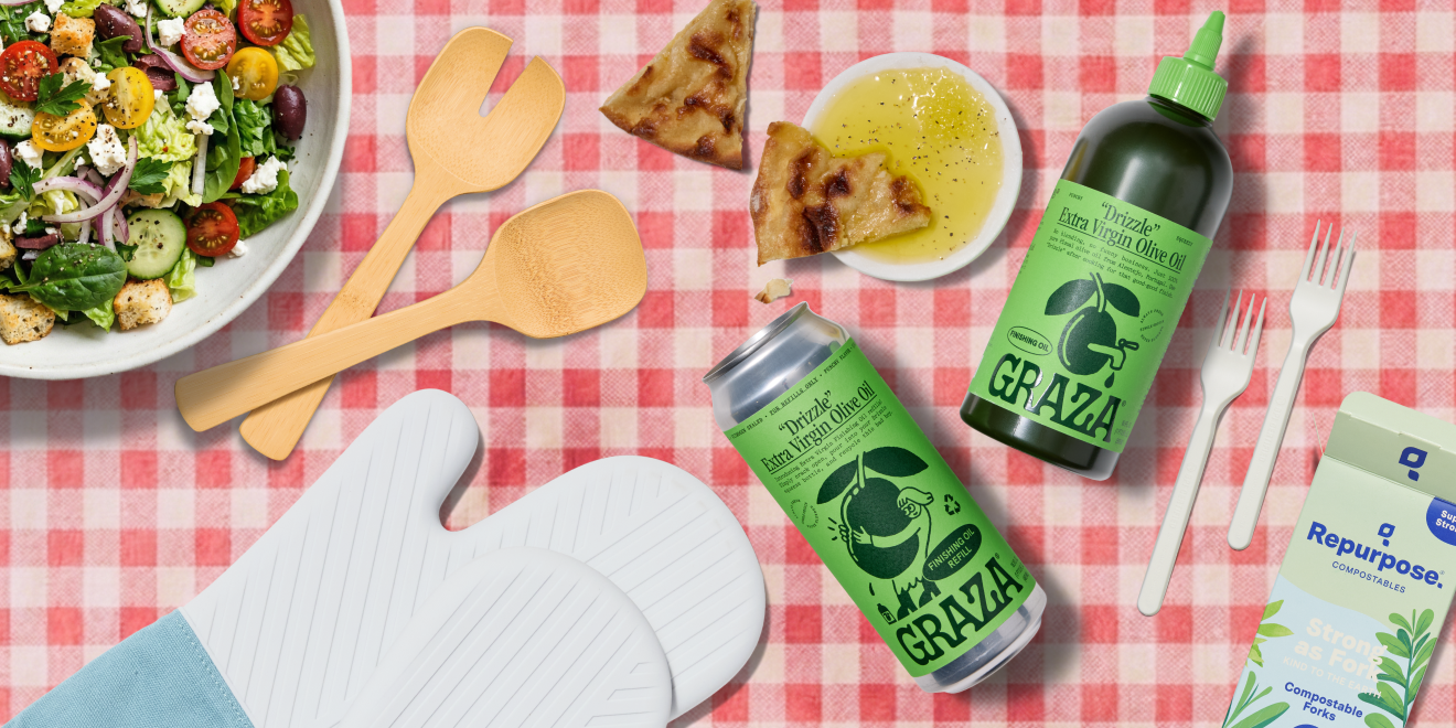 A picnic table with a salad bowl, wooden utensils, pita bread with olive oil, oven mitts, Graza olive oil bottle and can, and a box of Repurpose compostable straws on a red checkered tablecloth.