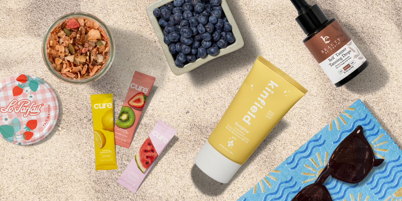 Healthy snacks, skincare products, a bottle of serum, a tube of sunscreen, sunglasses, and a bowl of blueberries arranged on sandy beach with a blue patterned towel.