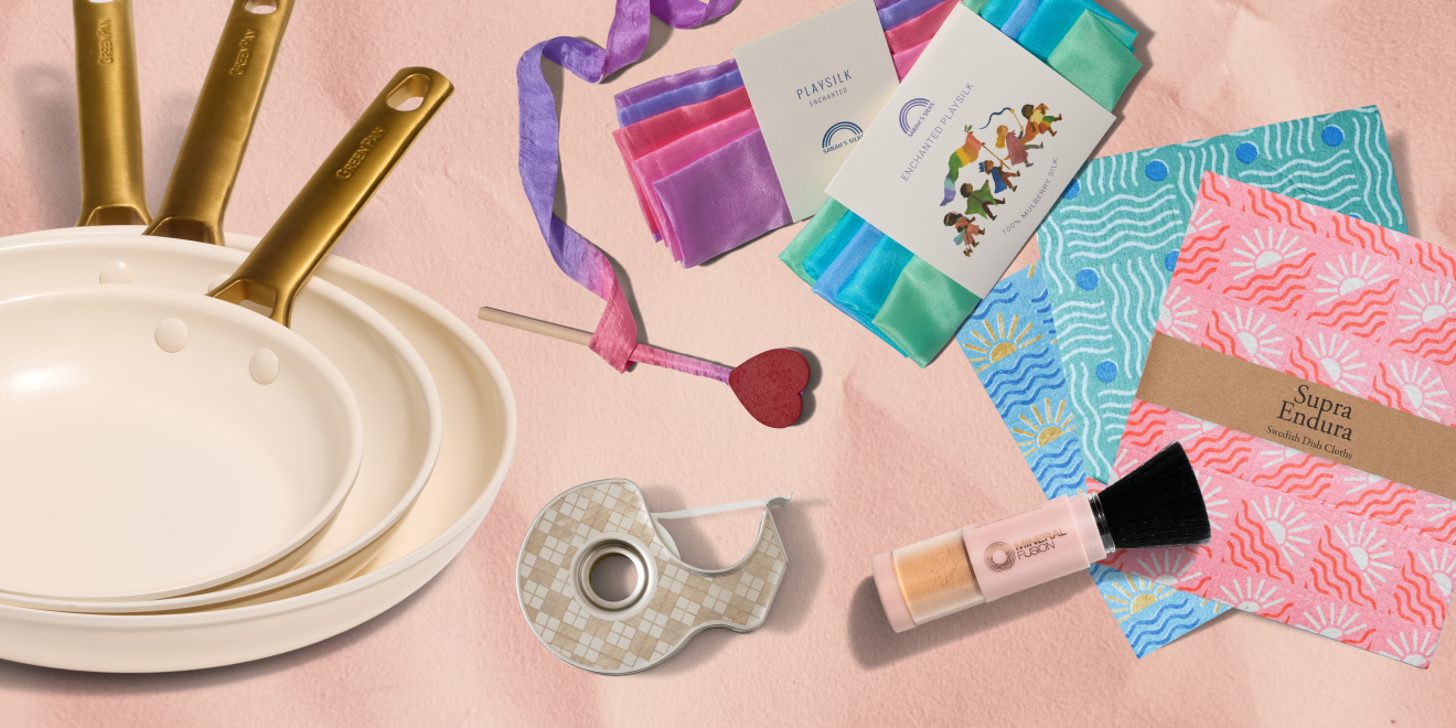 Three cream-colored frying pans with gold handles, colorful folded cloths, a heart-topped stick, patterned napkins, a roll of tape, and a makeup brush on a soft pink background.