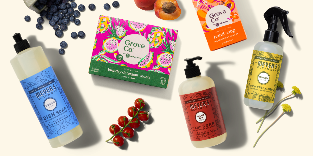 Assorted Mrs. Meyer’s and Grove Co. cleaning products arranged with fresh berries, tomatoes, apricots, and flowers on a light background. Products include dish soap, hand soap, air freshener, and detergent sheets.