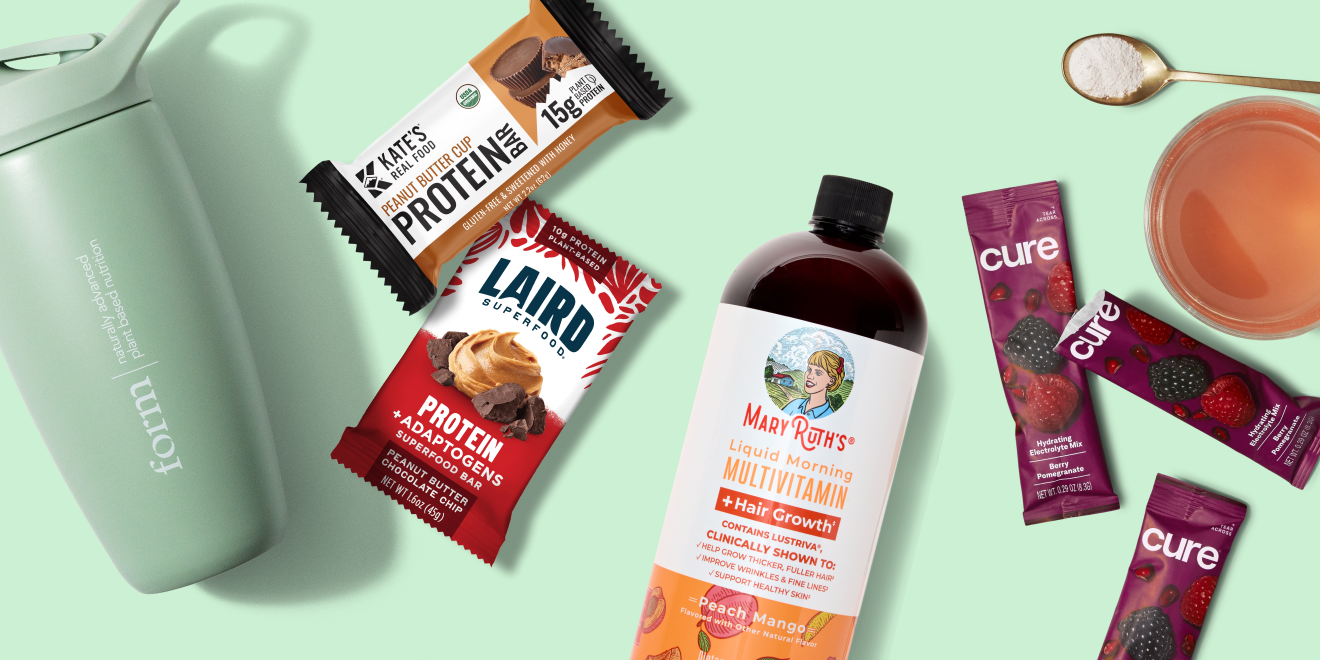 A green water bottle, two protein bar packs, a bottle of liquid multivitamin, two berry drink packets, a spoon with powder, and a glass of pink drink on a light green background.