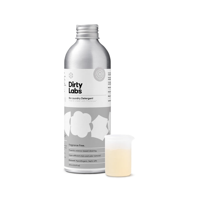 A silver bottle of Dirty Labs Bio Laundry Detergent with Phytolase® - Free & Clear, a fragrance-free laundry detergent ideal for sensitive skin, stands next to a small, clear measuring cup filled with light yellow liquid. The bottle features a minimalist white and gray label design.