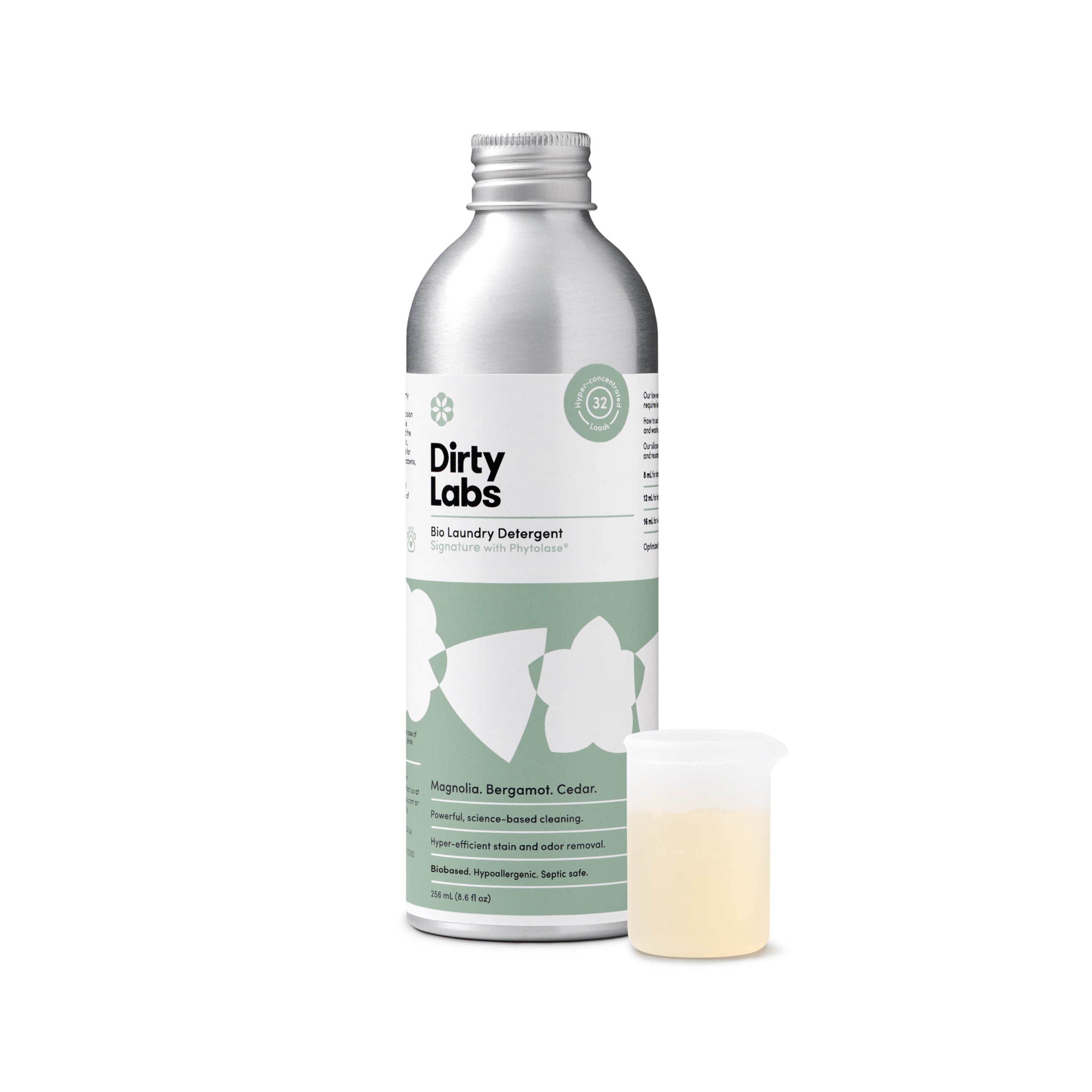 A silver bottle of Dirty Labs Bio Laundry Detergent with Phytolase® - Magnolia, Bergamot, Cedar stands upright beside a small measuring cup filled with light yellow liquid. The green and white label features floral graphics and product details, emphasizing its reputation as an eco-friendly, natural laundry detergent.