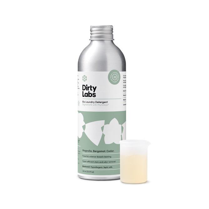 A silver bottle of Dirty Labs Bio Laundry Detergent with Phytolase® - Magnolia, Bergamot, Cedar stands upright beside a small measuring cup filled with light yellow liquid. The green and white label features floral graphics and product details, emphasizing its reputation as an eco-friendly, natural laundry detergent.