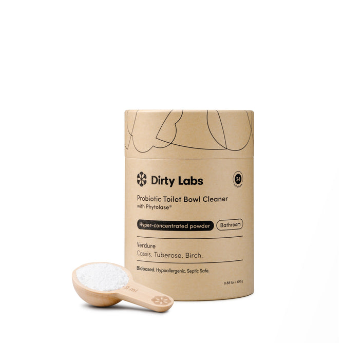 A cardboard container of Dirty Labs Probiotic Toilet Bowl Cleaner with Phytolase® is displayed beside a wooden scoop filled with white cleaning powder on a white background, emphasizing its powerful toilet bowl cleaning capabilities.
