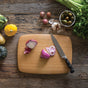 The Classic Bamboo Cutting Board by Bambu features a halved red onion, onion slices, and a knife on top, surrounded by tomatoes, garlic, radishes, herbs, yellow squash, and greens on a rustic wooden table.