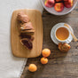 On a rustic table, the Bambu Classic Bamboo Cutting Board holds sliced bread—one with jam. Nearby sit apricots, a cup of tea with a pastry, and a bowl of peaches. A light cloth runs alongside this inviting arrangement.