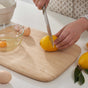 A person slices a lemon on the Bambu Classic Bamboo Cutting Board beside a glass bowl containing an egg and eggshells, with another lemon and some leaves close by.