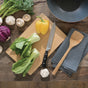 The Bambu Undercut Bamboo Cutting Board holds bok choy, a yellow bell pepper, and a knife on a table. Nearby are a striped cloth, wooden spatula, mushrooms, garlic, cabbage, and a black bowl.