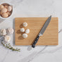 The Bambu Undercut Bamboo Cutting Board, paired with a chef's knife and five white mushrooms, is surrounded by a bowl of eggs, two garlic bulbs, and thyme on a marble countertop.