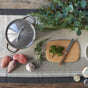 A pot with a lid, sweet potatoes, garlic bulb, fresh herbs, and Bambu’s Classic Bamboo Cutting Board with parsley and a knife are arranged on a striped tablecloth among green foliage.