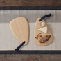 Two Bambu Artisan Droplet Cutting & Serving Boards, FSC-certified bamboo, rest on a striped placemat; one is empty, the other holds cheese, bread, a small container, and nuts. Both boards feature dark fabric handle loops.