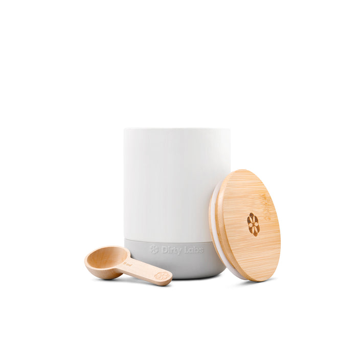 A white Refillable Ceramic Canister with a gray base, labeled Dirty Labs, is shown with a round bamboo lid and small scoop featuring a floral logo. All items are set against a white background.