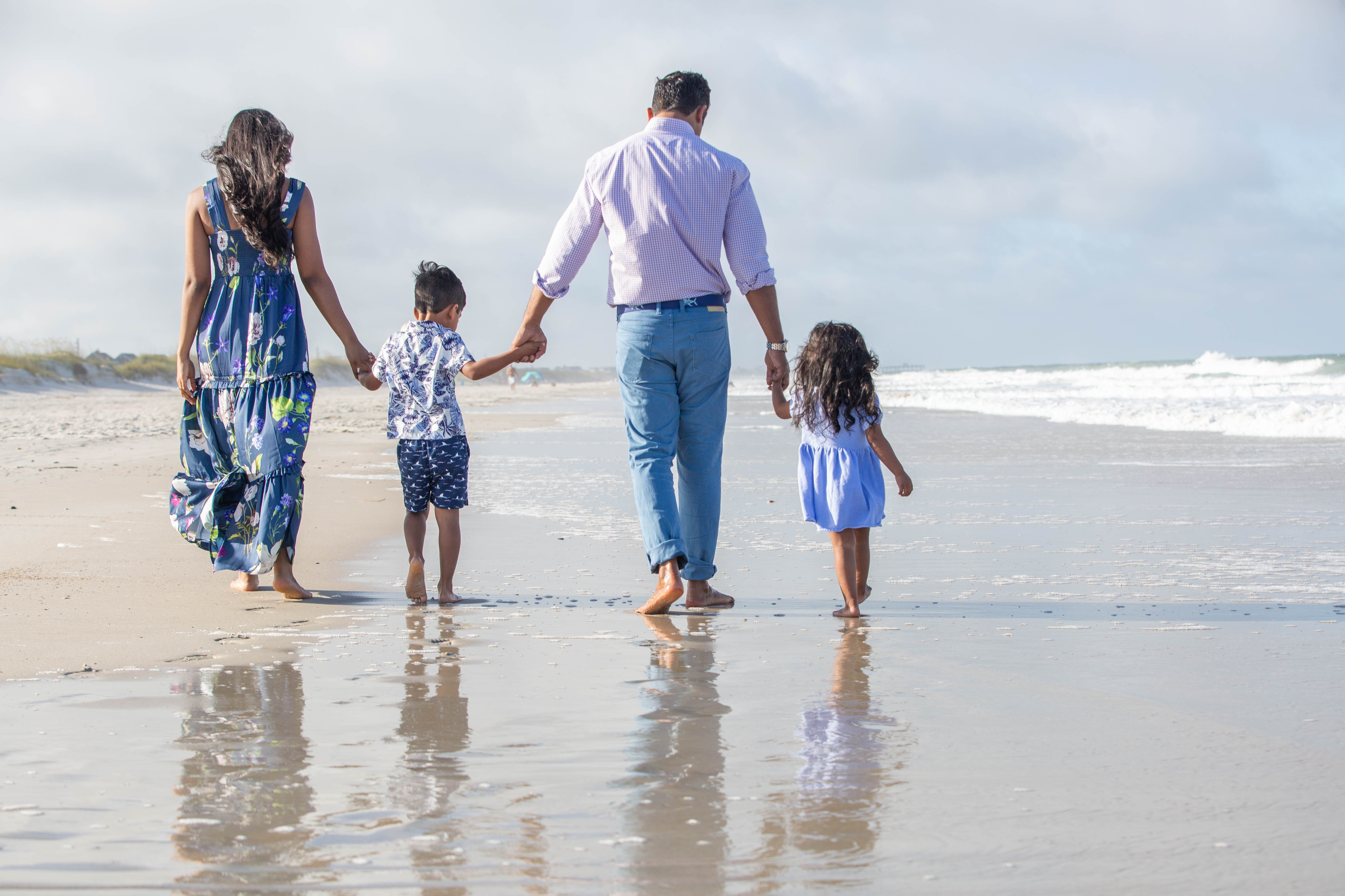A family of four, two adults and two children, walk hand-in-hand along a sandy beach near the waters edge, with their backs to the camera and their reflections visible on the wet sand.