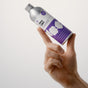 A hand holds up a silver bottle labeled Dirty Labs Bio Laundry Detergent with Phytolase® – Jasmine, Matcha, Vetiver, featuring a purple and white design, against a plain, light background.