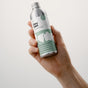 A hand holds a metallic bottle labeled Dirty Labs Bio Laundry Detergent with Phytolase® – Magnolia, Bergamot, Cedar, an eco-friendly laundry detergent featuring a green and white design, set against a plain light background.