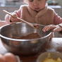 A young child uses the Bambu Kids in the Kitchen Set of 3 Bamboo Utensils (3Y+) to stir a bowl of brown baking mix, focusing intently while eggs and yellow batter rest on the table nearby.