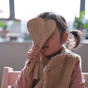 A young child playfully covers their face with the Bambu Kids in the Kitchen Set of 3 Bamboo Utensils (3Y+), wearing a pink top and beige vest, with blurred kitchen items and plants in the background.