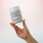 A hand holds up a white container of Dirty Labs 2-in-1 Bio Enzyme Dishwasher Detergent + Booster against a soft pink and white gradient background, showcasing the product’s powerful bioenzyme formula for an effective clean rinse.