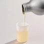 A metallic bottle of Dirty Labs Bio Laundry Detergent with Phytolase®—Jasmine, Matcha, Vetiver pours a viscous, pale yellow liquid into a small, translucent plastic measuring cup on a light surface.