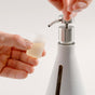 A hand dispenses liquid from a silver-top pump bottle of Dirty Labs Bio Laundry Detergent with Phytolase® - Free & Clear into a small, translucent measuring cup held by another hand. The bottle, labeled for sensitive skin, features a vertical window displaying the liquid level and a 300 mL marking.