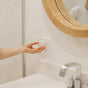 A person’s hand is plugging an Enviroscent Plug-in Air Freshener Refill into an electrical outlet on a white bathroom wall next to a mirror and a faucet.