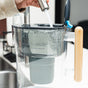 An individual holds the Epic Water Filters USA Nano XP Water Filter Pitcher | Reduces Bacteria & PFAS under a running faucet, filling it with water. The pitcher, with its distinctive wooden handle and NSF certified advanced filtration system, has its lid open as water splashes inside.