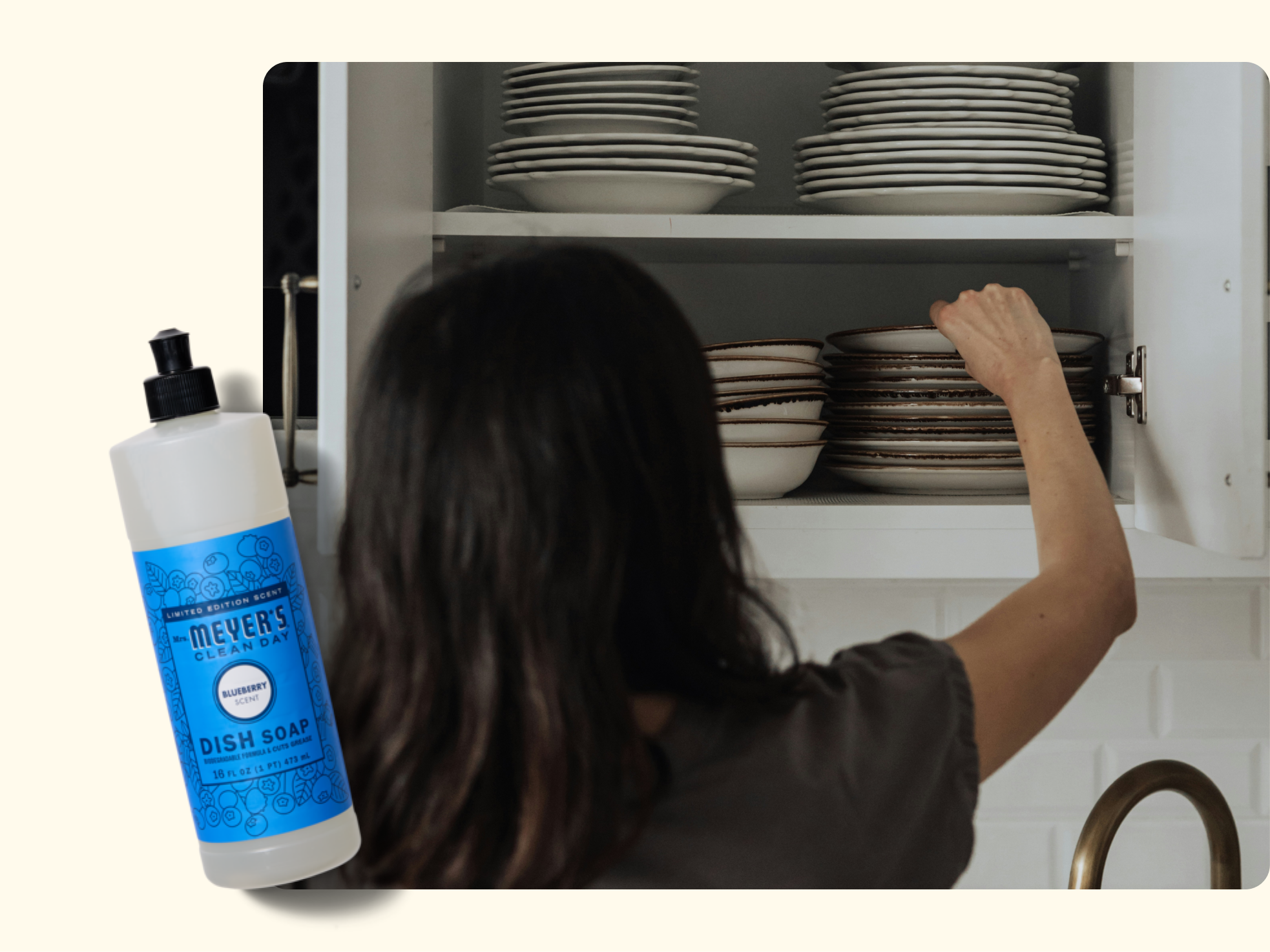 A person with long dark hair places a stack of bowls into a white kitchen cabinet filled with dishes. A bottle of Mrs. Meyers Clean Day dish soap is shown in the foreground on the left.