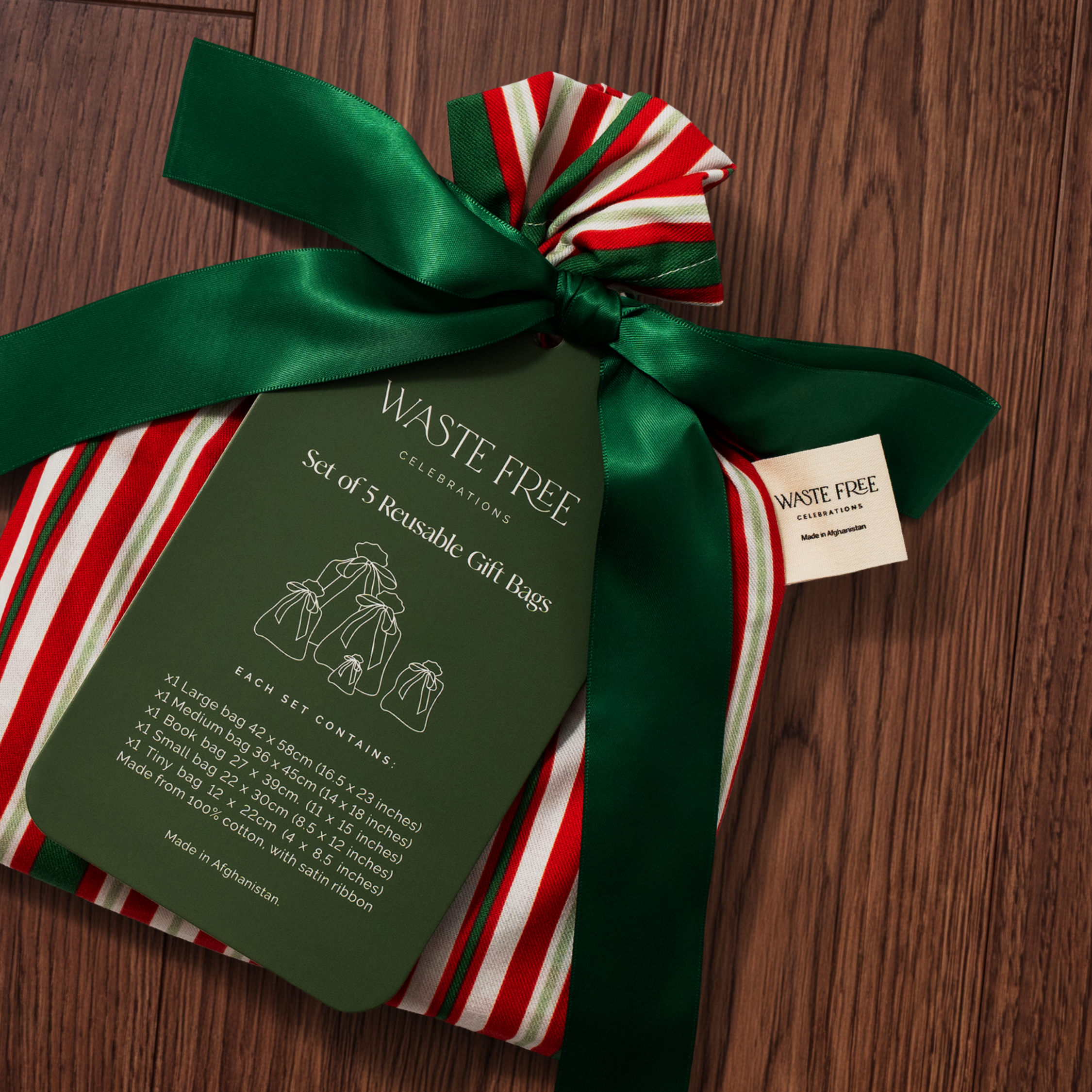 A wrapped gift set in a large green satin bow and red-and-white striped paper, with a hanging card reading 'WASTE FREE CELEBRATIONS Set of 5 Reusable Gift Bags.' The gift sits on a dark wood surface, and a small brand tag for Waste Free Collections is visible.