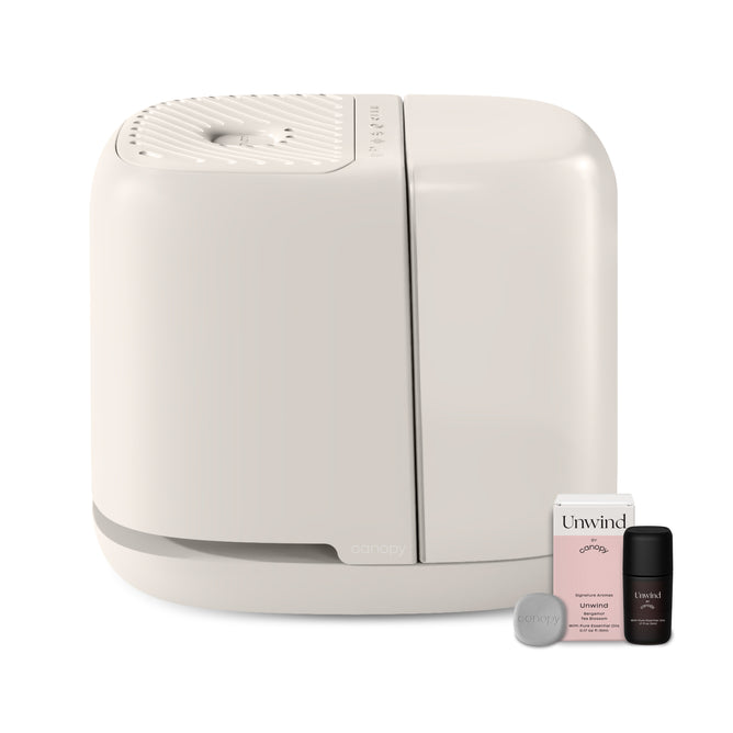 A modern white Canopy 2.0 Bedside Humidifier (with Filter) sits by an Unwind box, a small dark bottle, and a circular disk against a plain background. This dermatologist-recommended device uses mold-inhibiting technology.