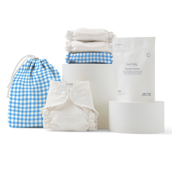 The Esembly Baby Cloth Diaper Try-It Kit, Size 1, is artfully arranged on white cylindrical stands with its blue and white gingham storage bag, folded cloth diapers, and packet of washing powder.