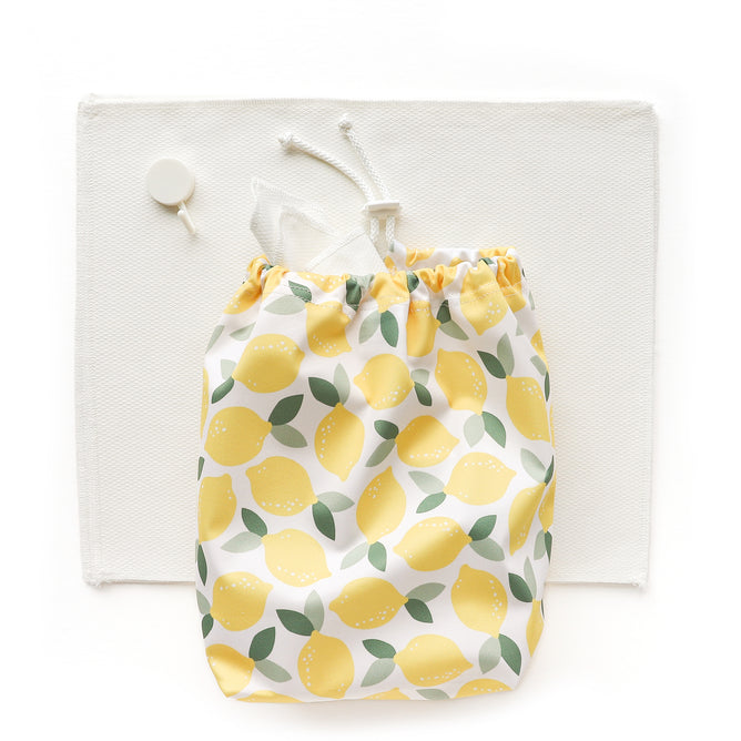 A white drawstring bag with yellow lemons and green leaves rests on a white placemat beside a small round object—ideal for storing your Esembly Baby Reusable Paper Towel Set (100% organic, unbleached).