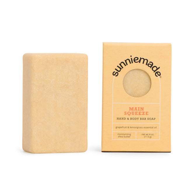 A rectangular bar of light beige soap rests beside its box labeled Sunniemade Moisturizing Hand & Body Bar Soap – Main Squeeze, featuring a round window that reveals the soap within. Infused with refreshing grapefruit and lemongrass, this bar is perfect for natural body care, delivering a gentle, moisturizing cleanse every time.