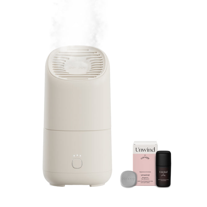 A Canopy Portable Humidifier releases mist beside a gray disk, an Unwind box, and a small dark bottle on a plain white background—ideal for hydrating your skin anywhere.
