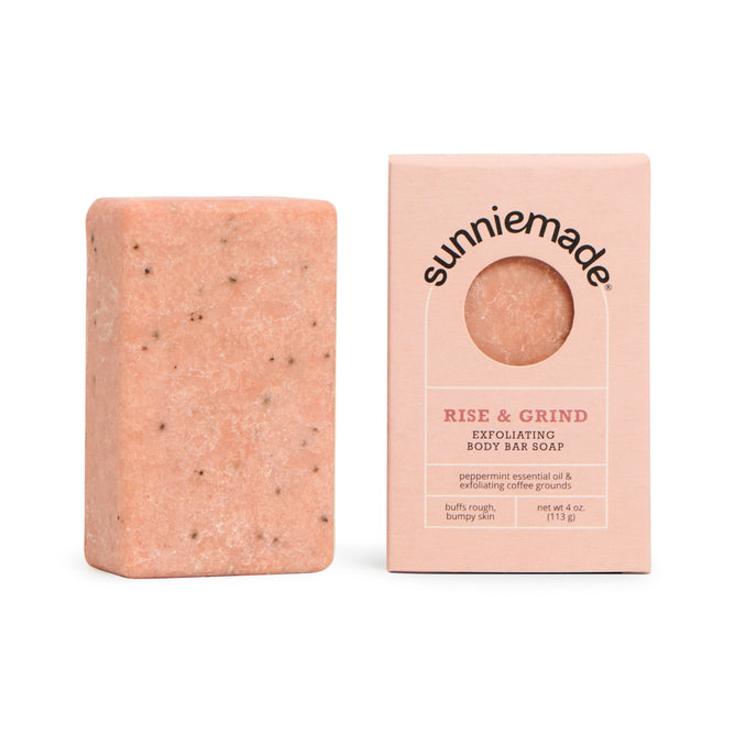 A pink rectangular Exfoliating Body Bar Soap - Rise & Grind by Sunniemade, flecked with dark coffee grounds for natural exfoliation, rests beside its box. The packaging features a round cutout displaying the soap inside and is labeled with the Sunniemade brand and product name, highlighting this unique body bar’s refreshing, coffee-infused cleanse.