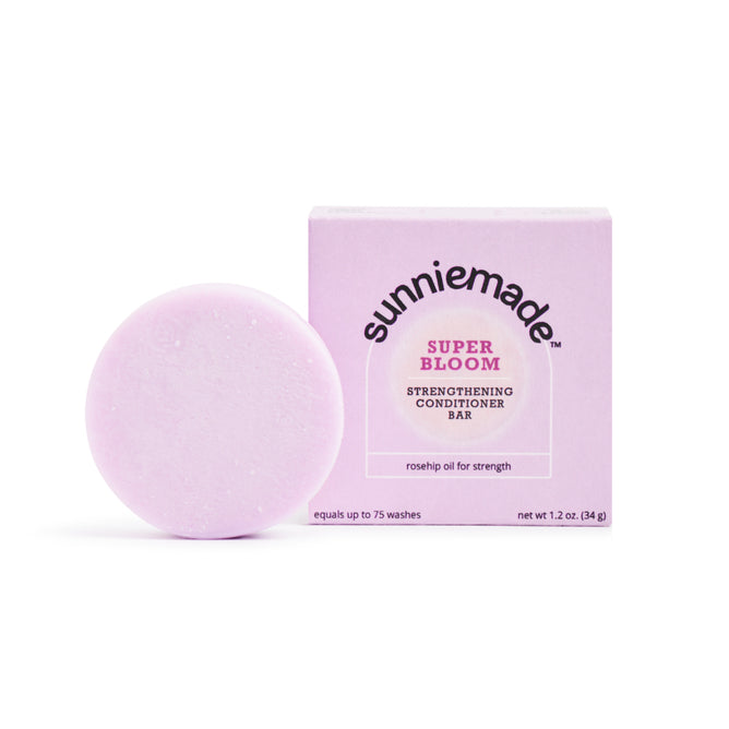 A round, light purple plastic-free Sunniemade Strengthening Conditioner Bar - Super Bloom sits next to its matching box featuring rosehip oil, on a white background.