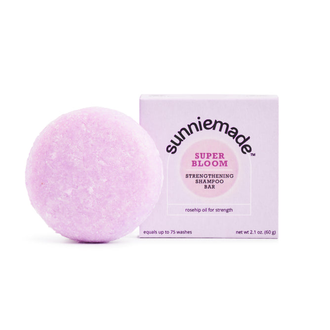 A round, light purple Strengthening Shampoo Bar - Super Bloom by Sunniemade is positioned next to its box, which reads “Sunniemade Strengthening Shampoo Bar - Super Bloom” and highlights rosehip oil for strength. Both the plastic-free bar and its packaging are displayed against a white background.