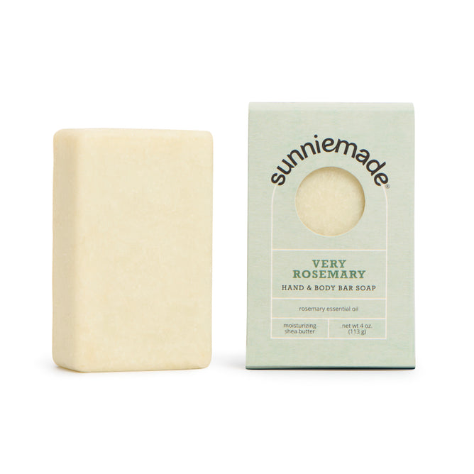 A rectangular bar of Sunniemade Moisturizing Hand & Body Bar Soap - Very Rosemary rests beside its box, which highlights its blend of rosemary essential oil, French green clay, and shea butter. The light green box, ideal for natural body care lovers, features a circular window.