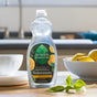 A bottle of Seventh Generation Dish Soap sits on a kitchen counter with fresh basil leaves and lemons, highlighting its dye-free formula and USDA-certified biobased status. A blurred sink and window appear in the background.