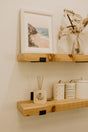Two wooden shelves display a beach photo in a white frame, a glass vase with dried flowers, an Enviroscent Stix Diffuser Air Freshener Refill for a non-toxic air freshening touch, and two white ceramic containers with lines and dots, all set against a beige wall.