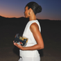 A woman in a sleeveless white outfit stands outdoors at dusk, holding The Parcel by Cadence. Her hair is styled in a neat bun as she faces left, silhouetted against a dark, mountainous background.