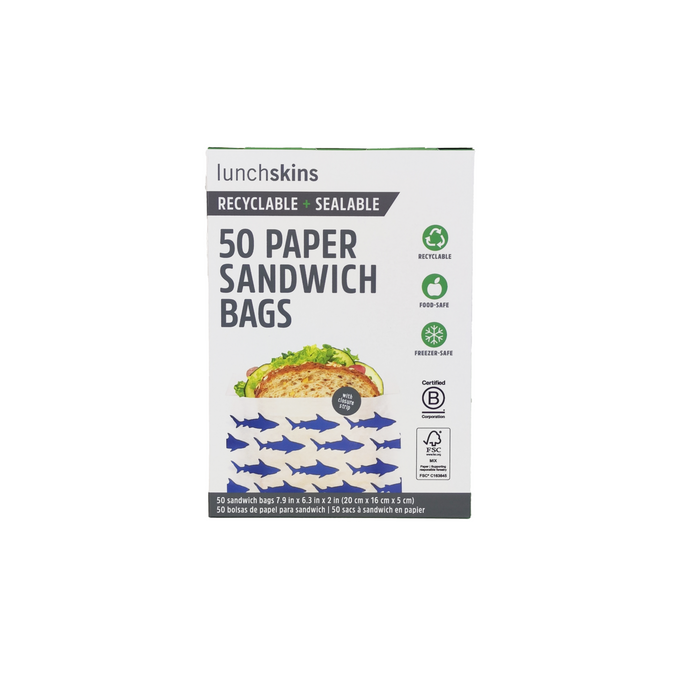 A box of Lunchskins Paper Sandwich Bags, adorned with a green leaf icon and blue shark illustrations, includes 50 eco-disposable bags. They are food safe, freezer safe, recyclable, sealable, and perfect for snacks and food storage.