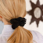 A person with long blonde hair in a low ponytail secured by a KOOSHOO Scrunchie, wearing a light gray top, is seen from behind. A star-shaped mirror hangs on the wall in the background.