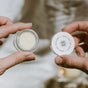 Two hands hold a small round jar of Honey Lip Treatment by Little Seed Farm with its white lid, which reads “GOOD FOR YOUR BODY | GOOD FOR THE EARTH.” The softly blurred background highlights the product.