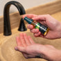 A person holding a small bottle of Badger Balm Organic Beard Oil - Soften & Nourish, dispensing it into their palm over a beige sink with a black faucet.