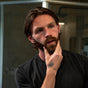 A bearded man with a tattooed hand stands in a modern bathroom, thoughtfully touching his chin as he looks in the mirror. Dressed in a dark shirt, he appears to be considering whether to use Badger Balm’s Organic Beard Oil - Soften & Nourish as part of his grooming routine.