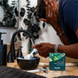 A person applies moisturizing shaving cream with a brush in front of a bathroom mirror. A razor, soap dispenser, and a box of Badger Balm Shave Bar - Smooth & Moisturize are on the counter next to a black bowl.